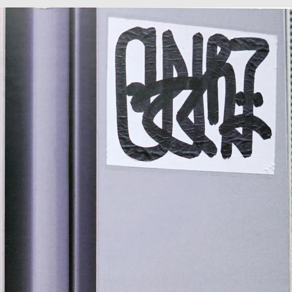 Close-up shot of a graffiti sticker with bold, black handstyle lettering on a white background, placed on an urban surface in Tokyo. A perfect snapshot of the city's sticker culture.